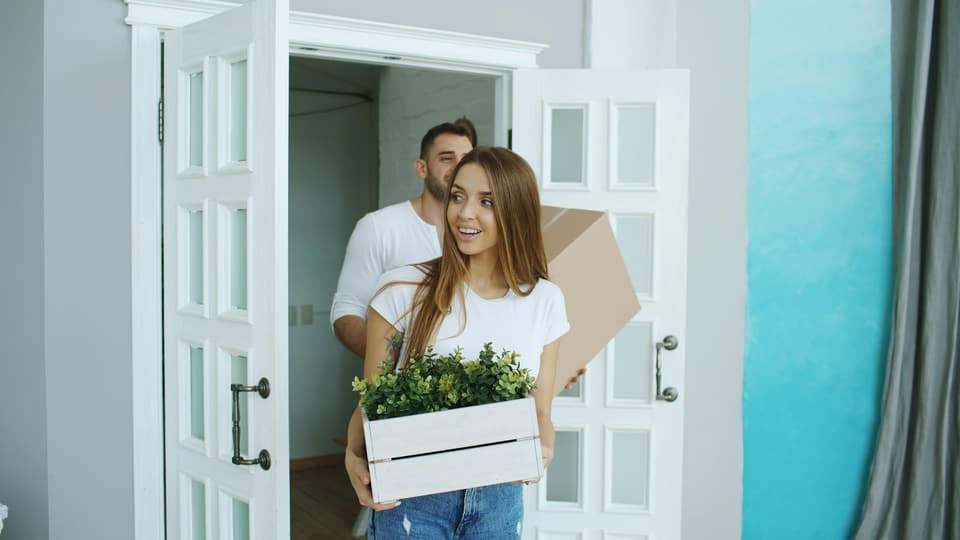 A couple carrying a moving box and a crate of plants through a doorway